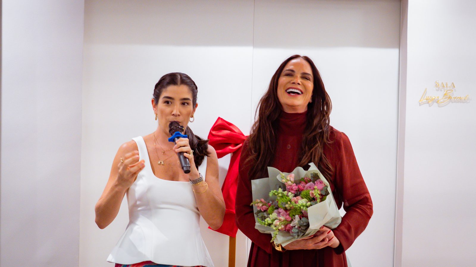 Camilla Liporaci, presidente do Instituto Nós Por Elas, discursa ao lado de Luiza Brunet diante da porta da sala que leva o nome da embaixadora, ainda com o laço vermelho da inauguração.