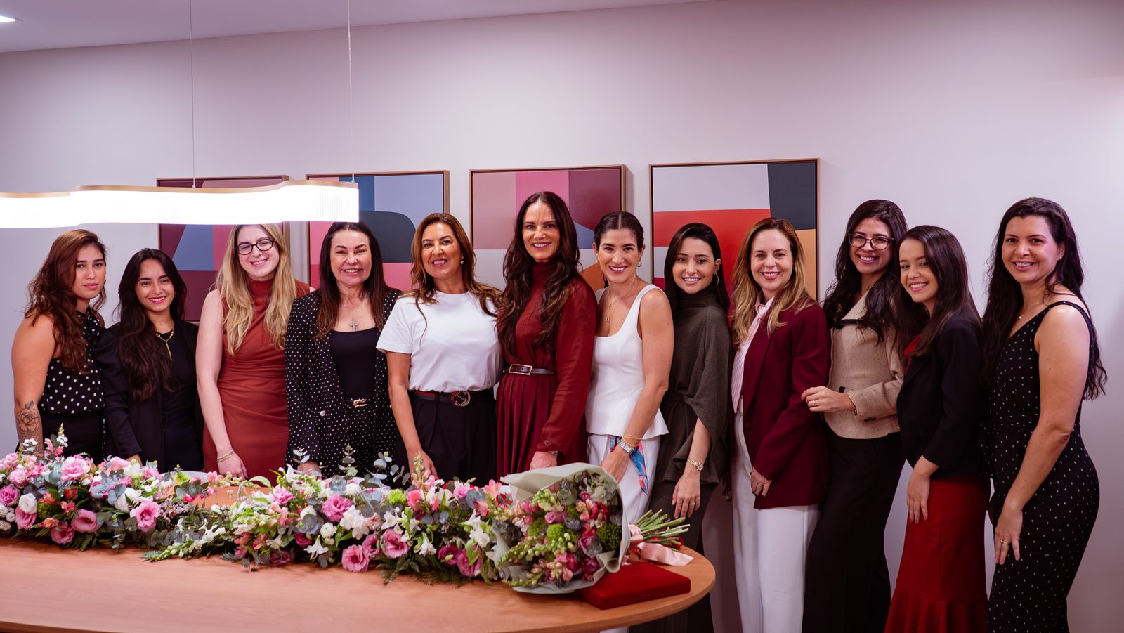 Luiza Brunet e convidadas posam juntas atrás de uma mesa com arranjos de flores, a placa e o buquê da homenagem.